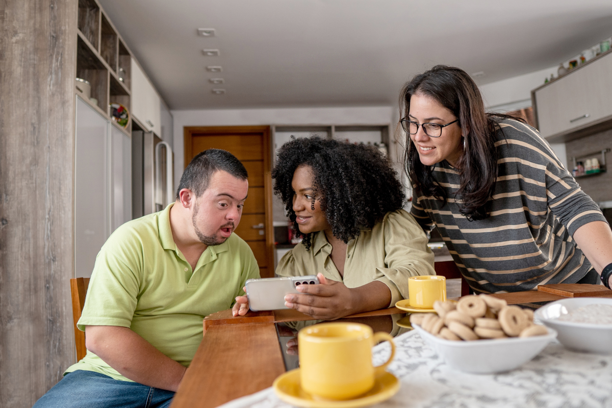 Three people, two women and a man, are sitting at a breakfast table looking at a phone together. There are mugs and a bowl of cookies on the table in a bright, modern kitchen setting.