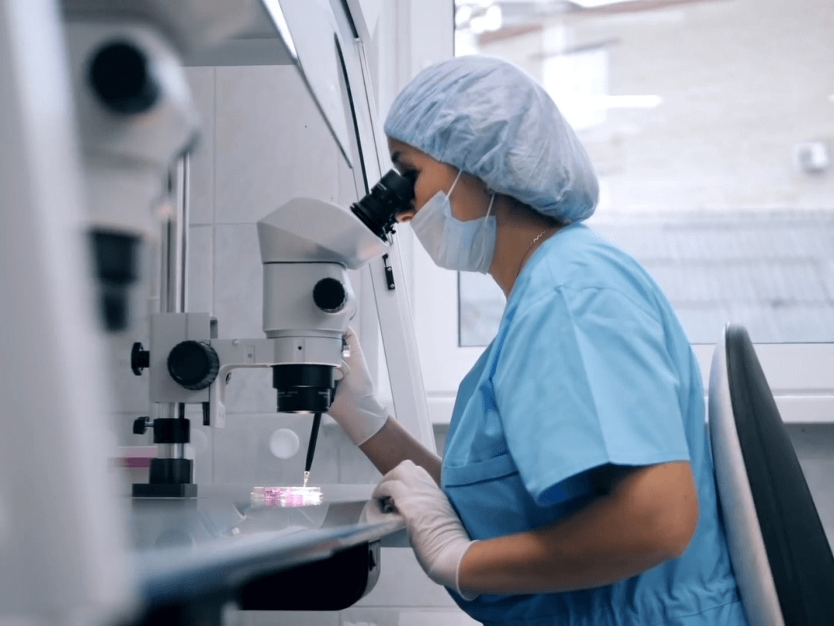A laboratory technician in scrubs looks through a microscope