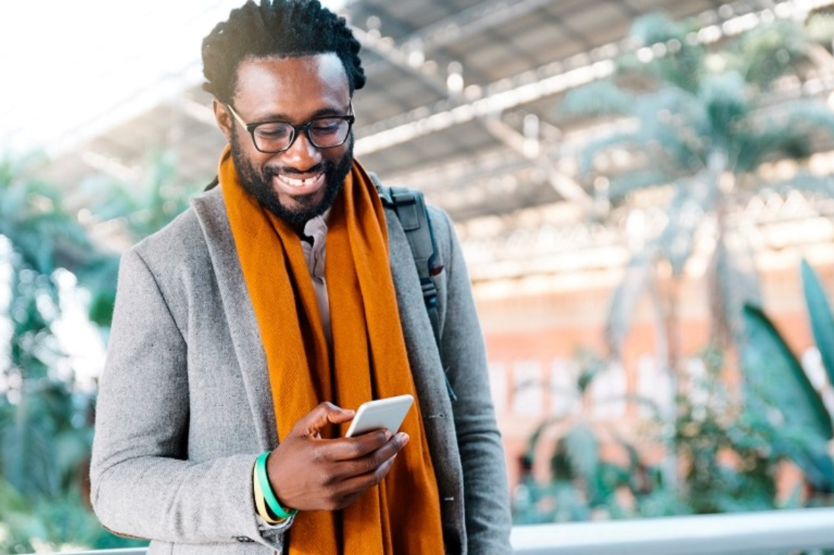 A person wearing a gray coat and orange scarf holds a smartphone in a bright, indoor setting with plants and a glass roof.
