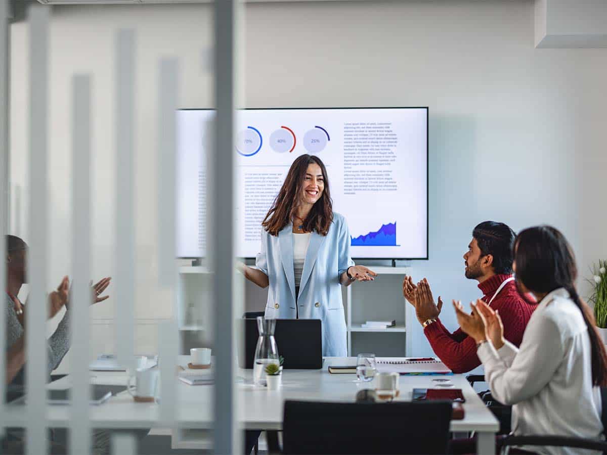 A woman stands in front of a screen giving a presentation in an office meeting room while attendees seated around a table clap and show appreciation. The screen displays graphs and charts.