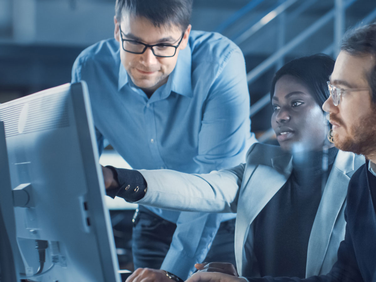 A diverse group of professionals collaborating and discussing work while looking at a computer monitor in a modern office environment, representing teamwork and innovation in artificial intelligence or machine learning.