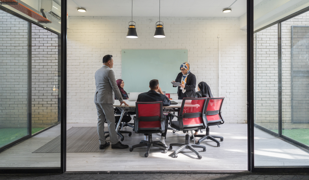 A group of professionals having a meeting in a modern office conference room with large glass walls, red chairs, and a white brick wall background.