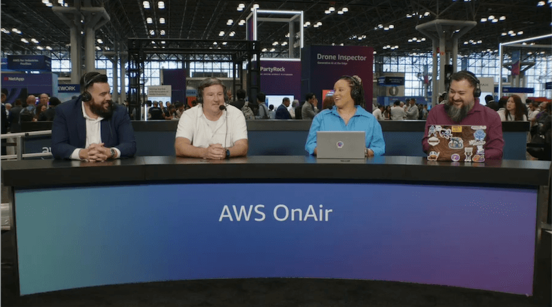 Alt-text: A panel table with "AWS OnAir" branding, featuring four individuals seated with laptops, in a busy conference setting.