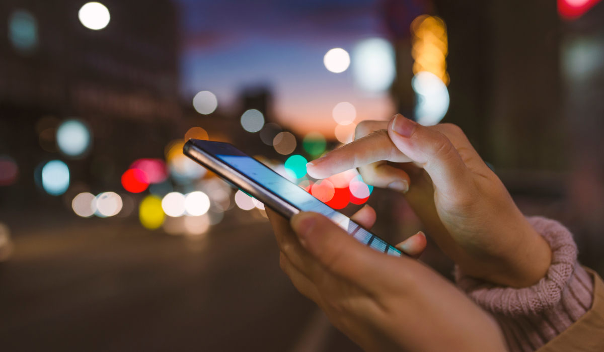 A close-up of hands using a smartphone at night in a city, representing generative AI's impact on the telecommunications industry and mobile technology.