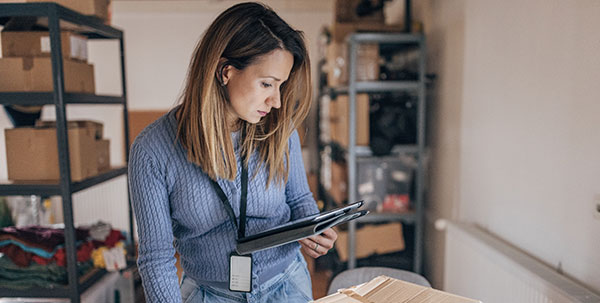 A woman conducting inventory management in a small business setting, examining products and using a tablet in a storeroom environment.