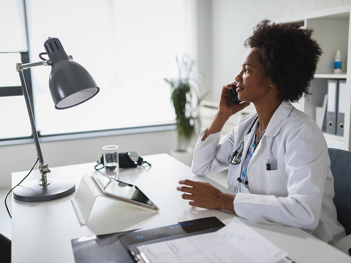 A doctor in a white coat with a stethoscope around their neck sits at a desk, talking on the phone, with a tablet, lamp, and documents on the desk.