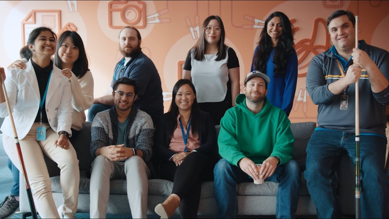 A group of people posing for a group photo in a casual office setting, sitting and standing together on and around a sofa, with a decorative wall in the background.