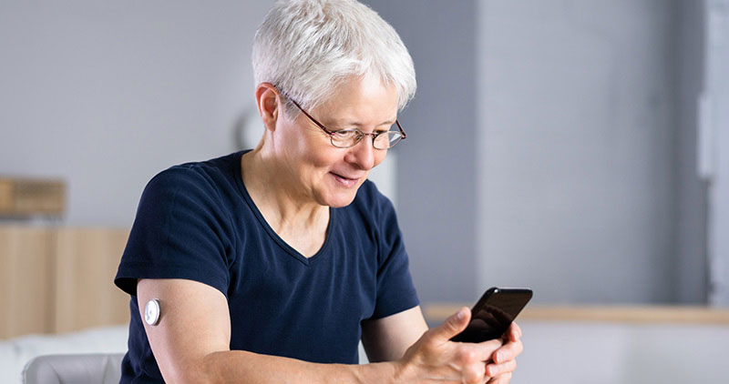A senior adult with short white hair, wearing glasses, is sitting indoors and smiling while using a smartphone. The person has a glucose monitoring sensor attached to their upper arm, indicating the use of digital health technology.