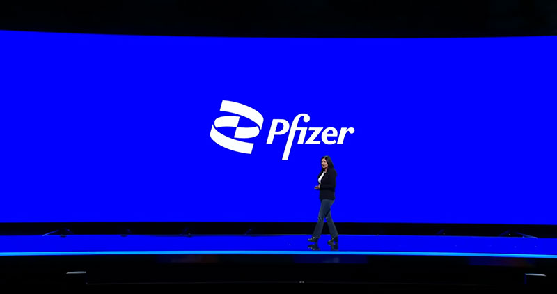 A person stands on stage in front of a large blue screen displaying the Pfizer logo.