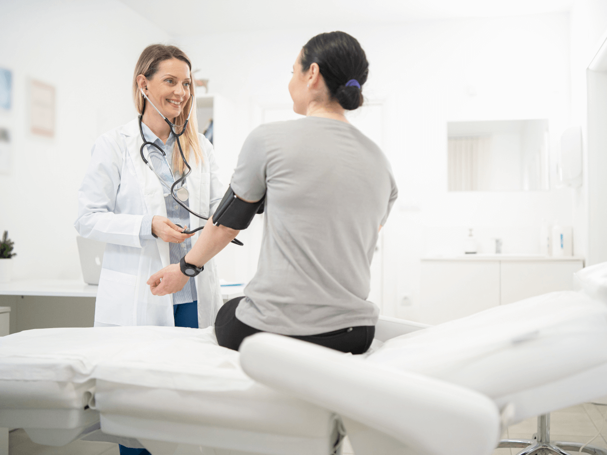 A doctor checks the blood pressure of a woman patient in a clinic setting. The scene depicts a healthcare professional providing care using a blood pressure cuff during a medical appointment.