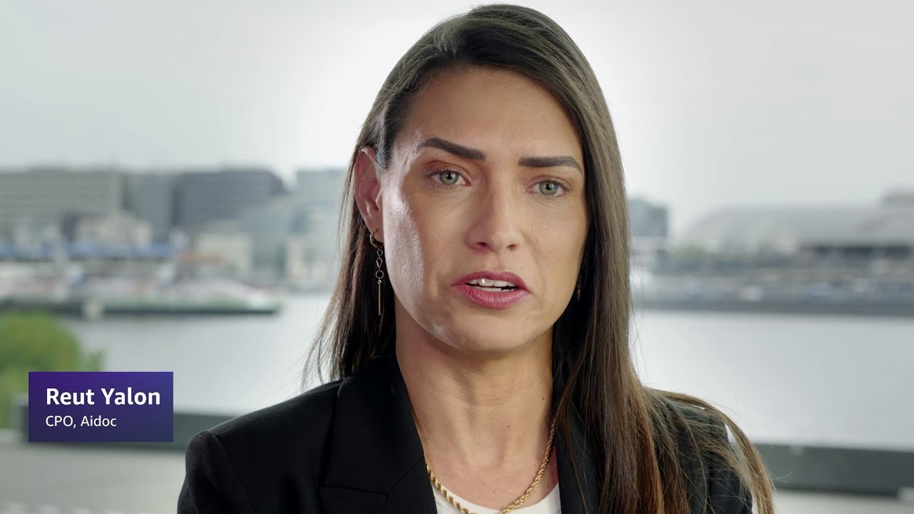 A woman with long brown hair wearing a black blazer and gold necklace, with a blurred cityscape and water in the background; text overlay reads "Reut Yalon, CPO, Aidoc."