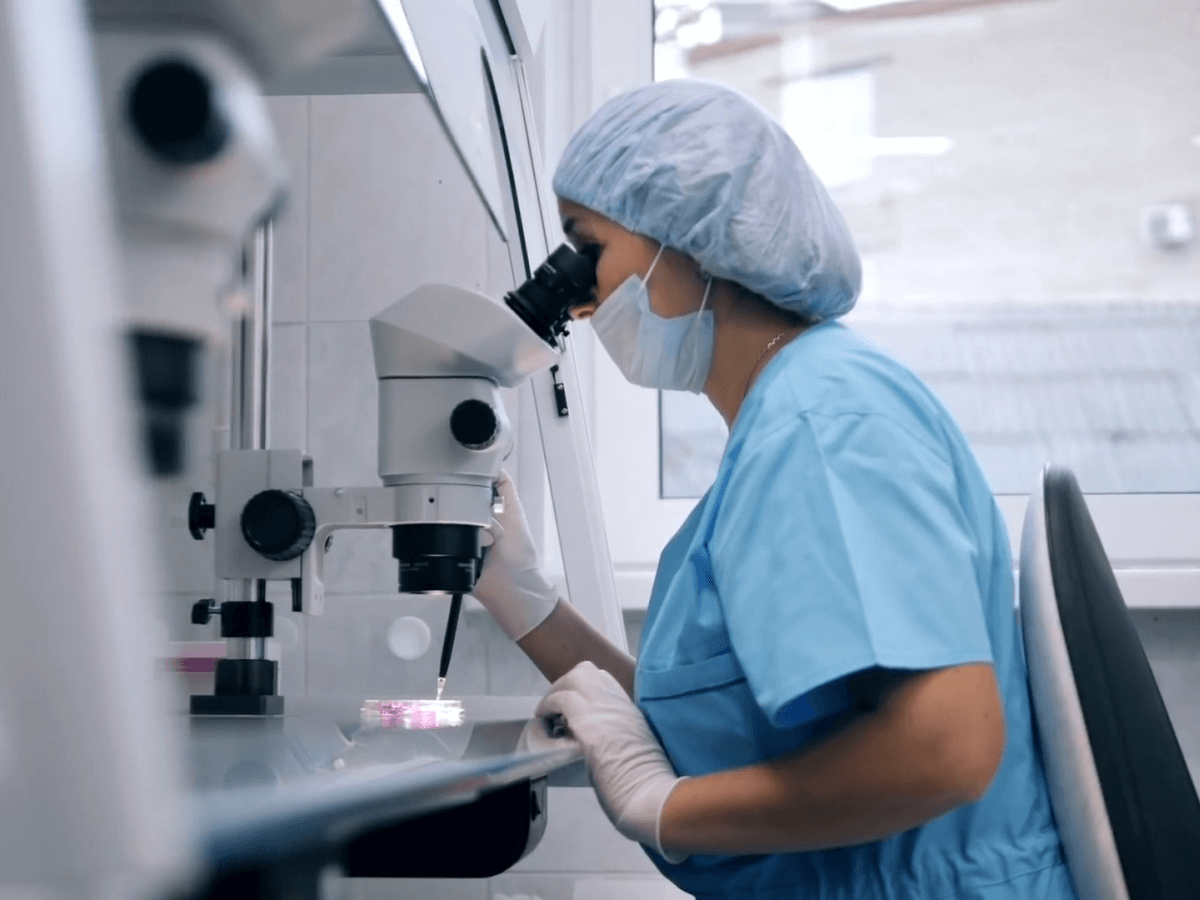 A doctor or scientist in scrubs, mask, and gloves using a microscope in a laboratory setting.