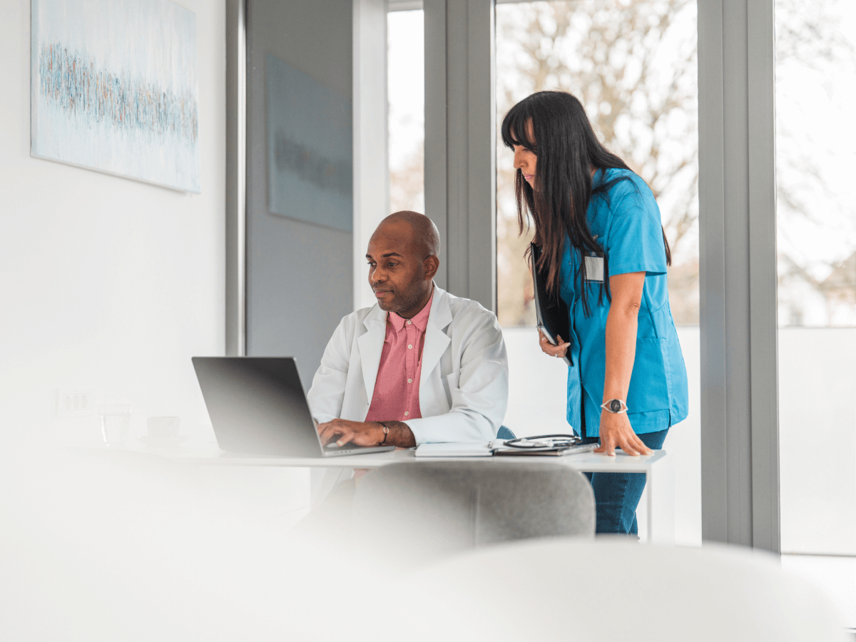 Two doctors looking at a computer screen together in a modern medical office setting. One is seated at the desk using a laptop while the other stands and looks on, both engaged in reviewing patient or medical data.