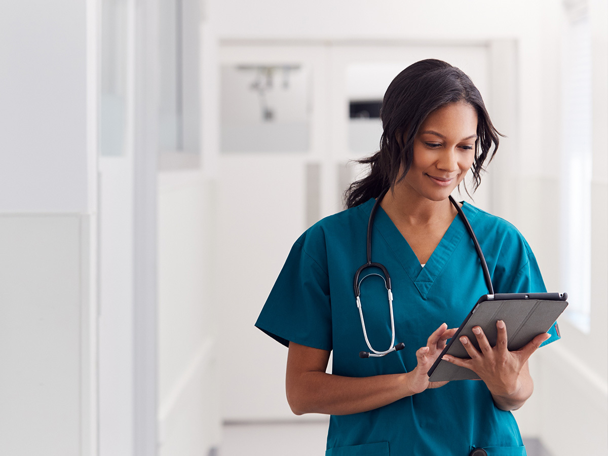 A healthcare worker in teal scrubs with a stethoscope around their neck, holding and using a tablet in a bright, clinical hallway.