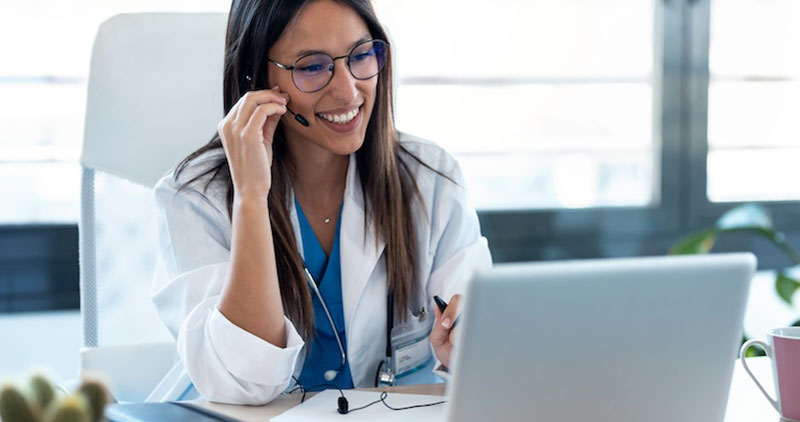 A doctor wearing a headset and white coat smiles while providing a telehealth consultation using a laptop in a modern office setting.