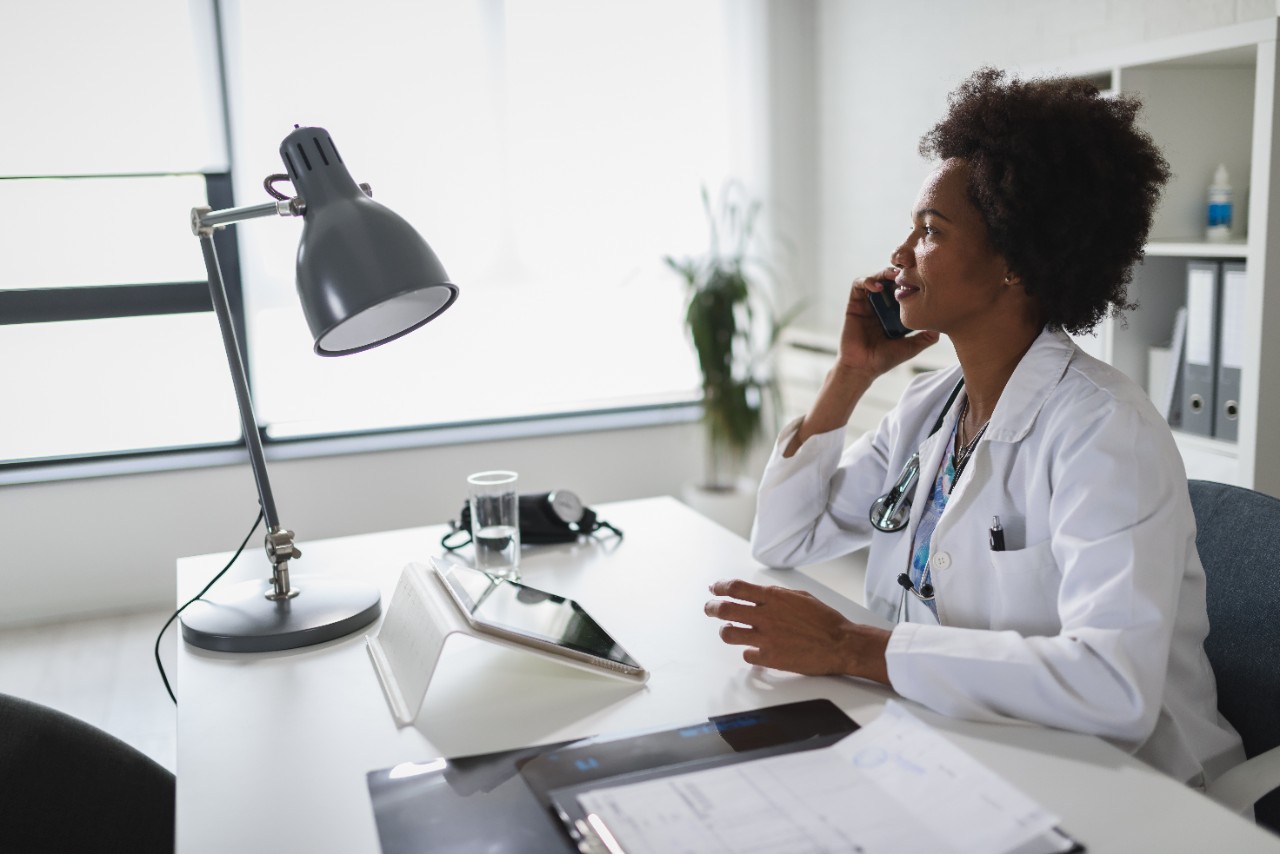 A clinician in a white coat sits at a bright office desk, taking a call on a mobile phone with a tablet and paperwork in front of her.