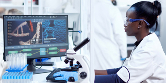 A medical technician wearing safety glasses works at a laboratory workstation with a computer displaying genomic analysis and a microscope, illustrating the use of technology in genomics research.