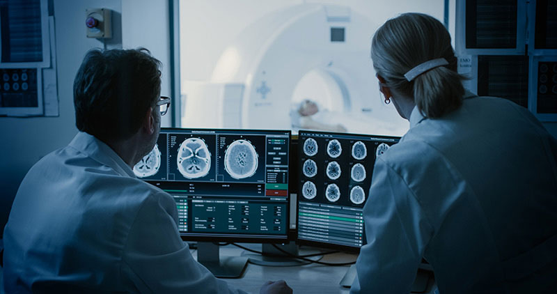 Two doctors review brain scan images and data on computer monitors while a patient is inside an MRI machine in a medical imaging room.