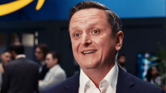 A business executive in a suit being interviewed at an automotive industry event, with a blurred background of people and an Amazon-themed setting. Represents the collaboration between Volkswagen and AWS for AI/ML solutions in the automotive sector.