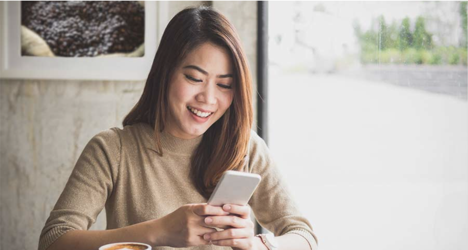 A person sitting at a cafe table smiling and looking at a smartphone, with a coffee cup in front and a window in the background.