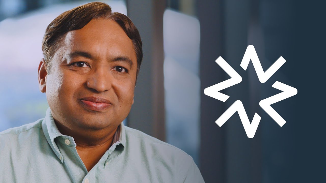 A man smiling in a professional setting with a modern geometric logo on the right side, suitable for use as a YouTube thumbnail related to AI, ML, or technology topics.