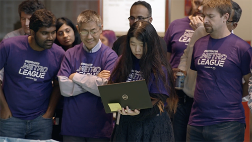 A group of people wearing purple "Metro League" shirts gathered around, with one person holding a laptop.