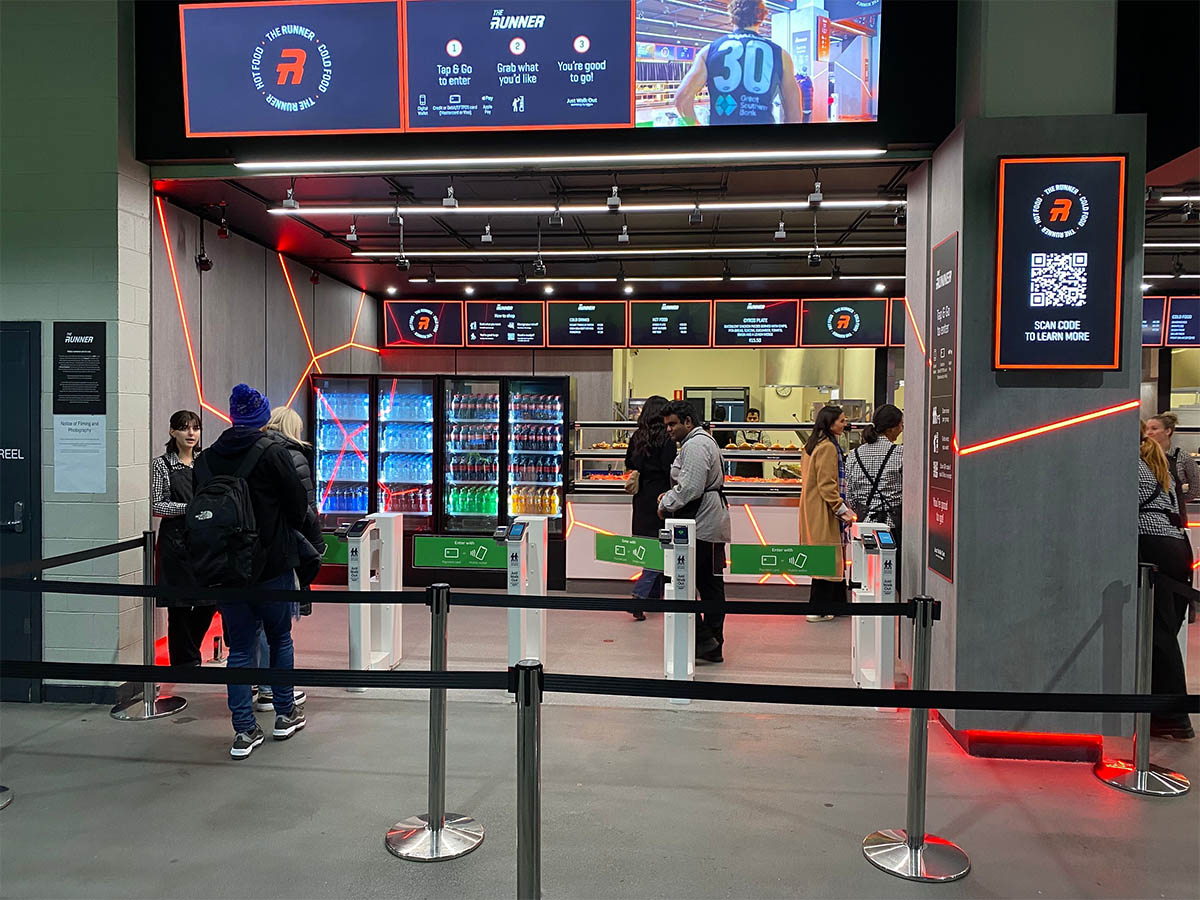 Customers enter a modern, AI-powered convenience store using automated gates, select food and beverages from refrigerators and counters, and exit without traditional checkout lines, demonstrating frictionless retail with machine learning technology.