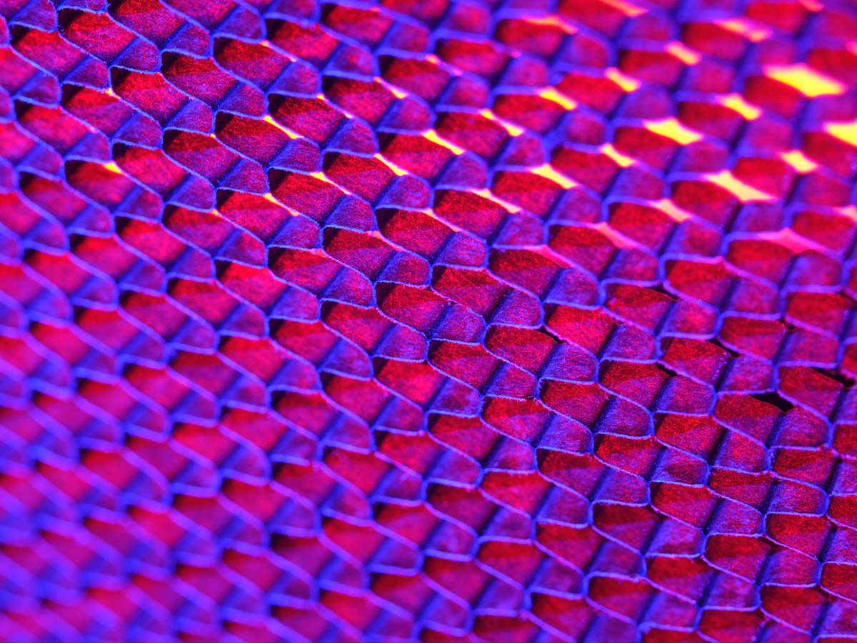 An abstract close-up image featuring a vibrant pattern of red and violet hues, resembling a geometric or honeycomb texture, with bright highlights and a modern, digital art aesthetic.