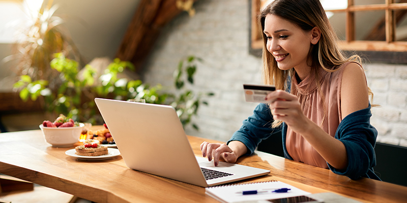 A woman sits at a table using a laptop and holding a credit card, likely making an online purchase. The setting is a cozy home environment with natural light and plants in the background.