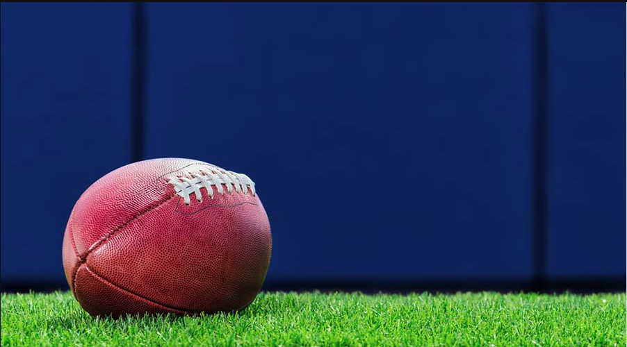 A close-up image of an American football resting on green grass with a blue background, representing the NFL.
