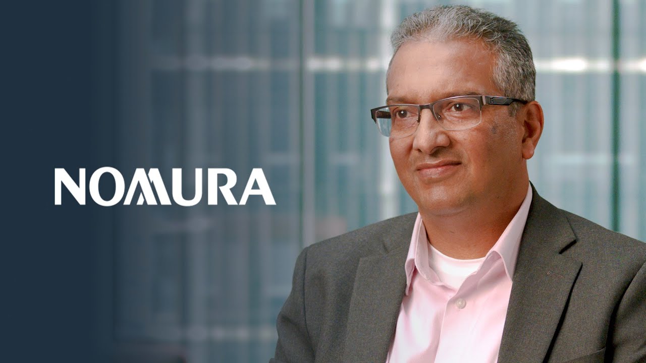 A business executive in a suit with glasses seated in front of an office background. The Nomura logo is prominently displayed to the left.