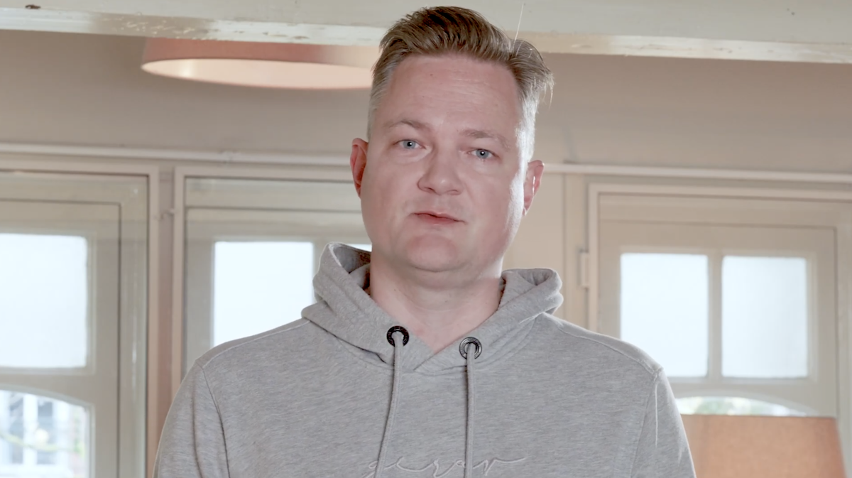 A person wearing a gray hoodie stands indoors with windows and soft lighting in the background.