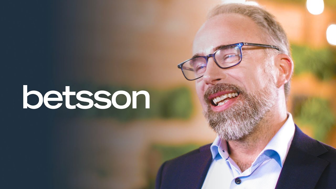 The Betsson logo displayed on a dark background with a blurred indoor setting featuring warm lighting and greenery.