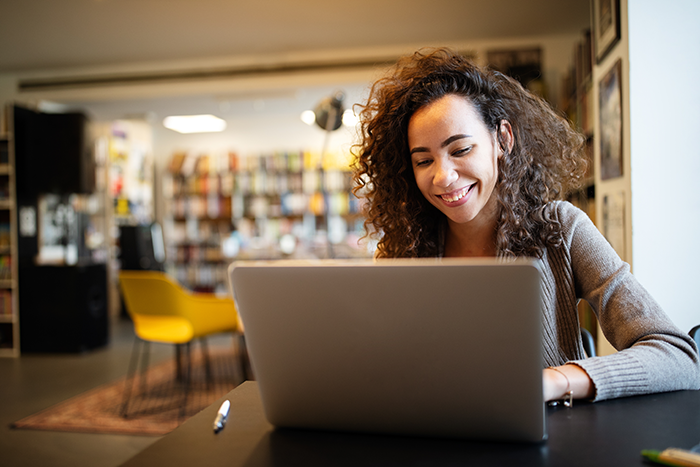 A woman sitting at a table in a library or bookstore, smiling while working on a laptop. The setting is modern and cozy with bookshelves and a yellow chair in the background.