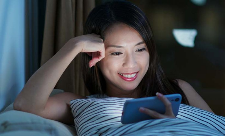 A woman is smiling while looking at her smartphone, lying on a couch in a relaxed home environment.