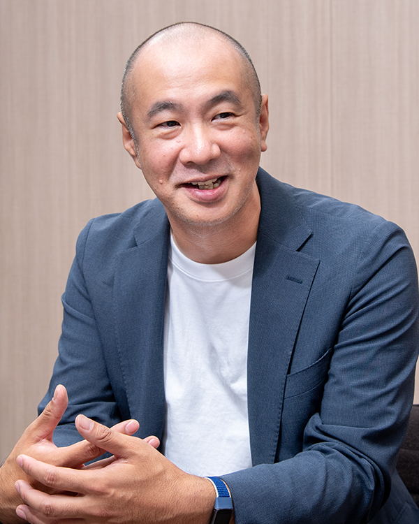 Portrait interview photo of a man in a blue jacket and white shirt, smiling and gesturing with his hands, seated against a neutral background.