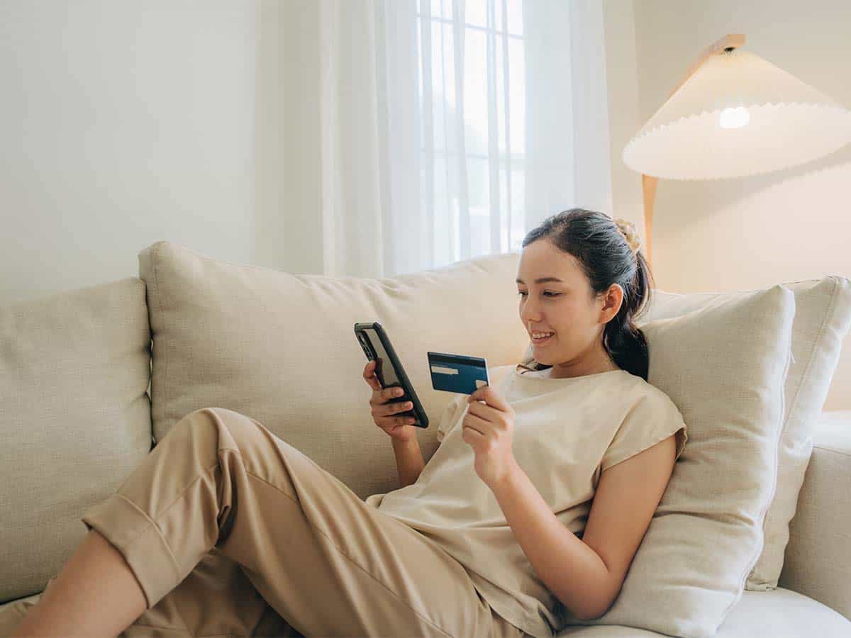 A woman is sitting on a couch, smiling while holding a credit card and using a smartphone, suggesting online shopping or digital payment.