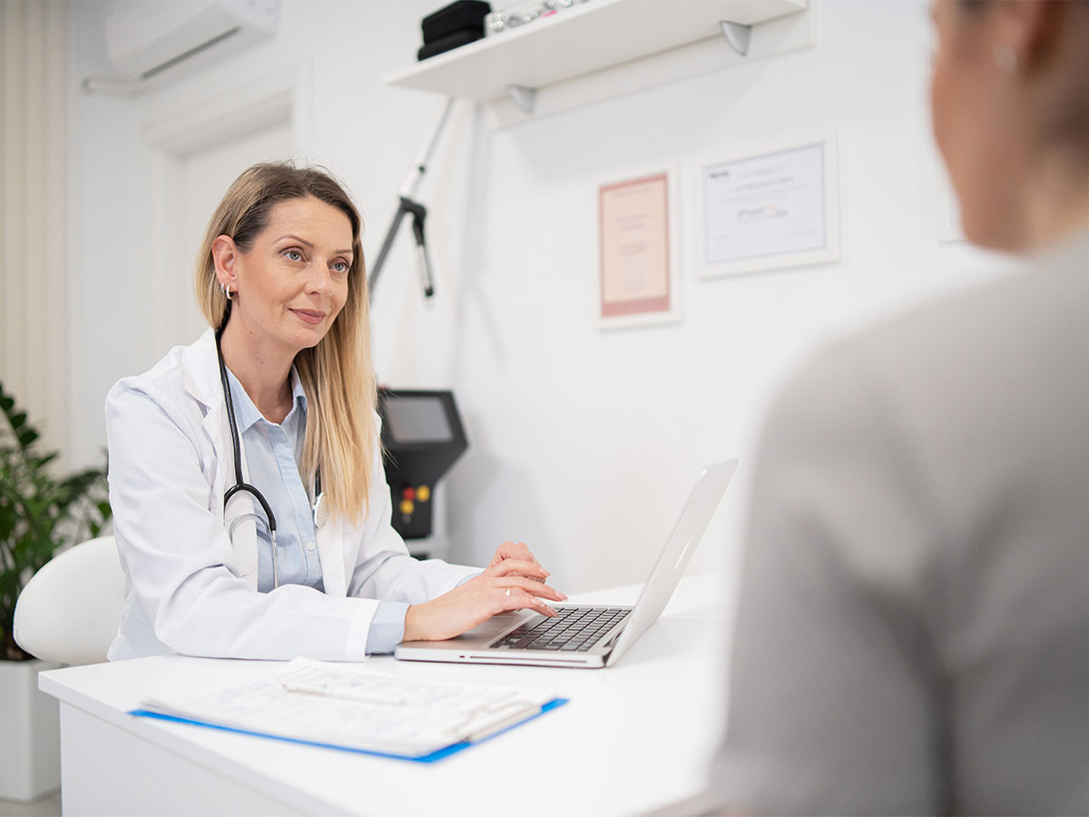 A doctor in a white coat with a stethoscope is sitting at a desk, speaking to a patient in a medical office. The doctor is using a laptop during the consultation.