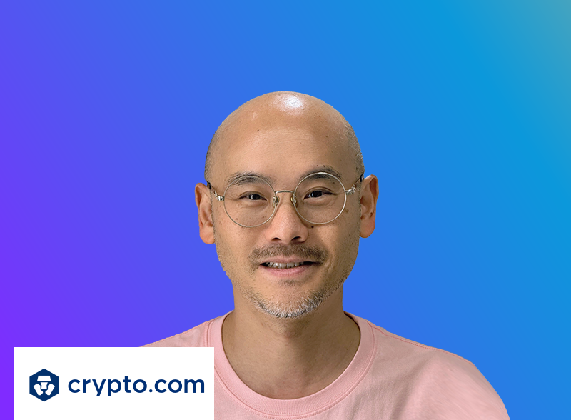 A person wearing glasses and a pink shirt is pictured in front of a gradient blue background with a 'crypto.com' logo and branding in the lower left corner.