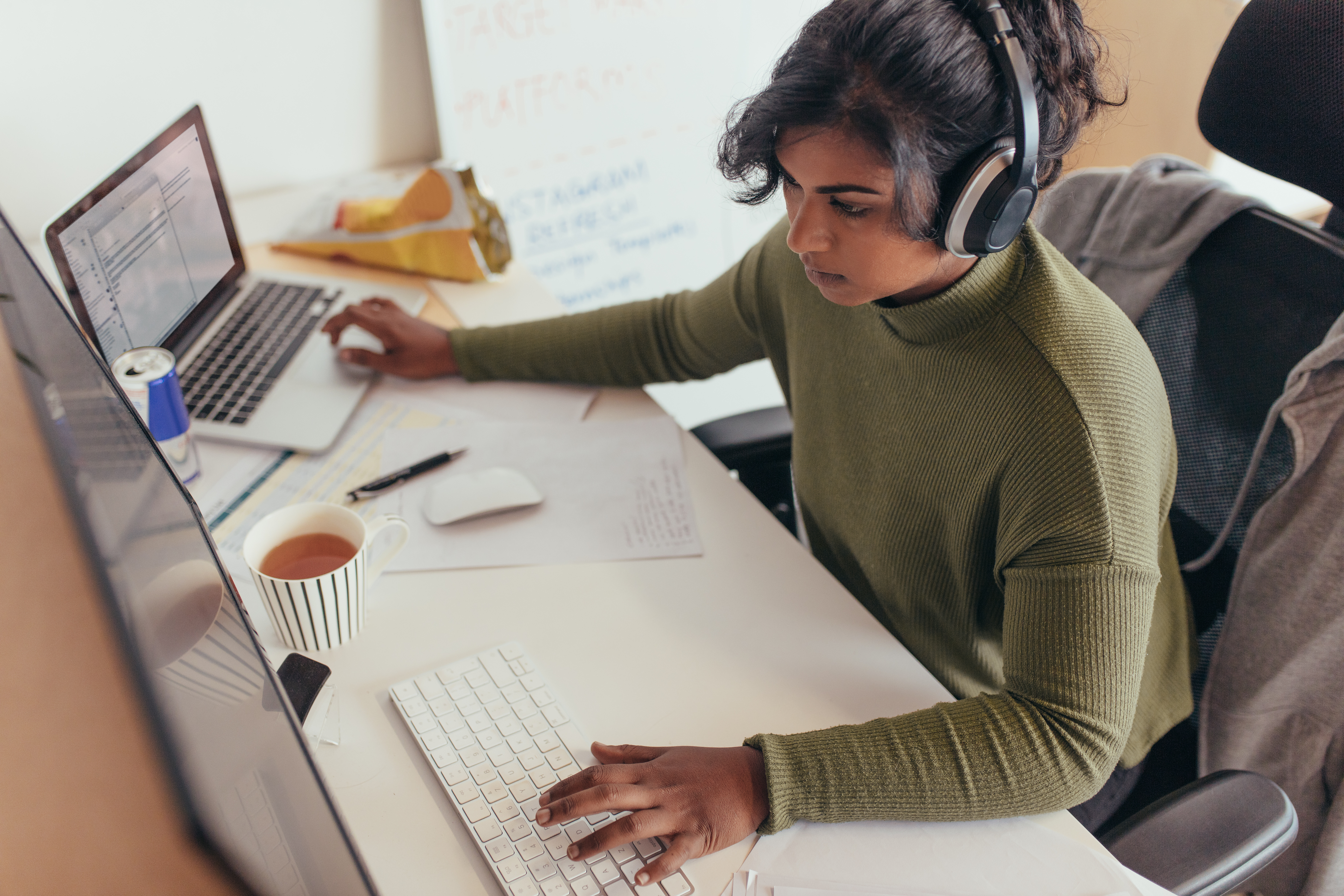 Female programmer coding on a desktop computer and laptop at her desk in office. Young woman in casuals wearing headphones working on a laptop and desktop computer.