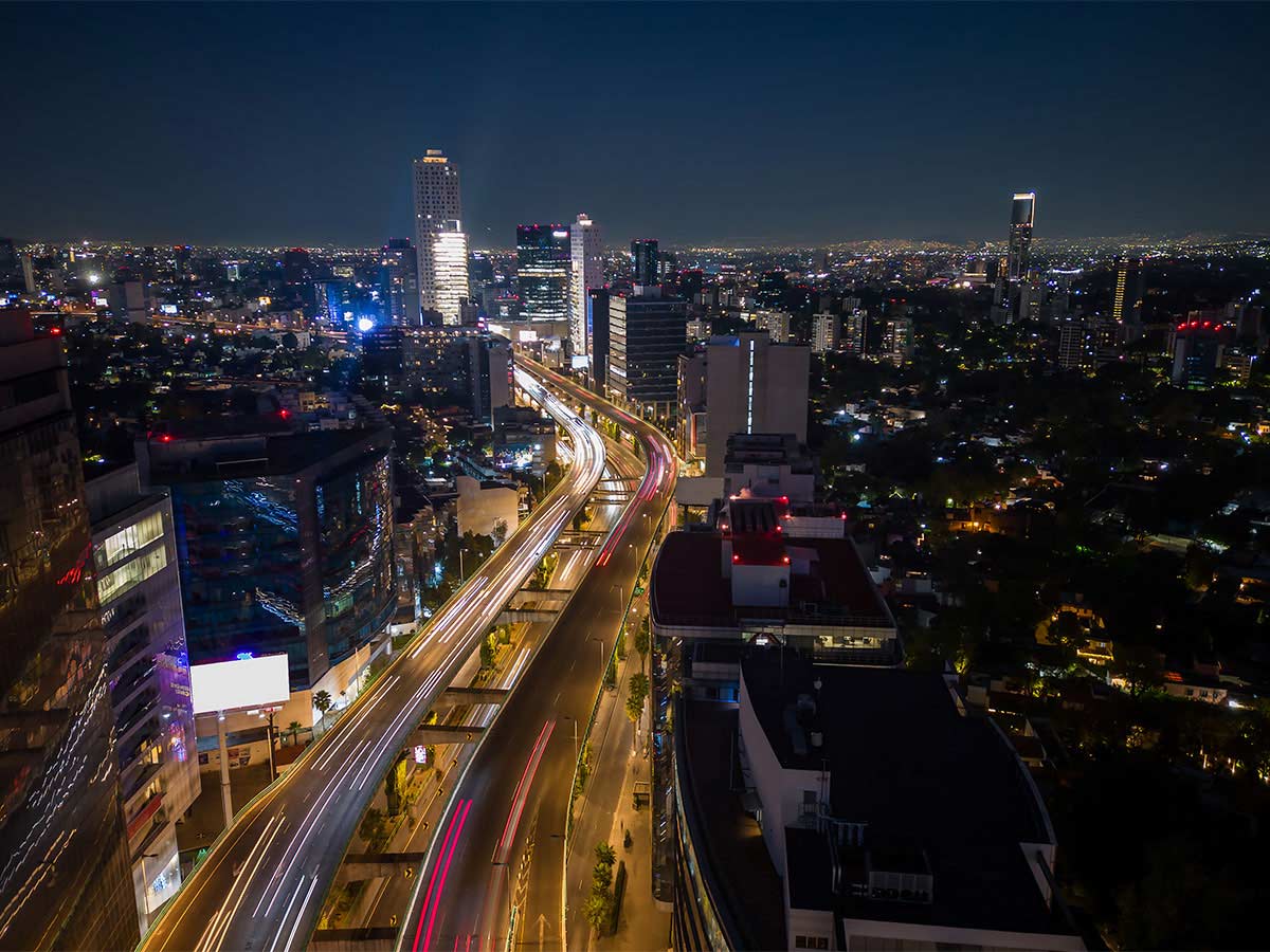 Aerial view of city roads illuminated at night, showing light trails from vehicles and city skyscrapers in the background.