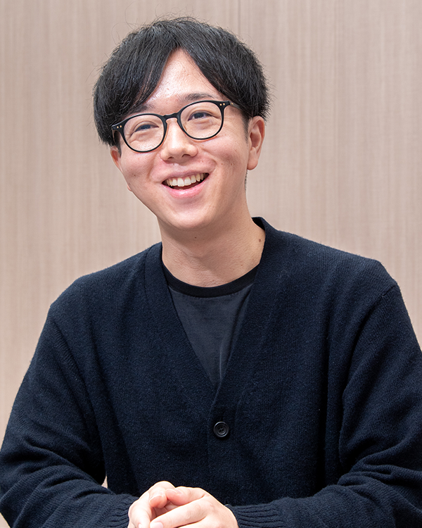 A smiling person with short dark hair and glasses, wearing a black sweater, sitting in an interview setting with a neutral background.