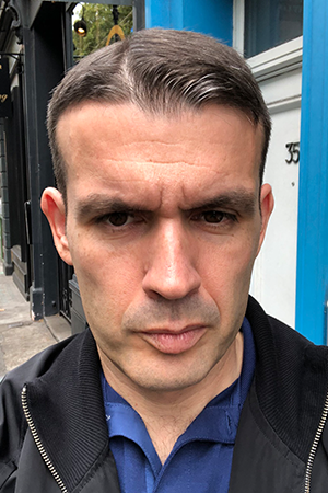 A serious-looking man takes a selfie outside near a blue and white building.