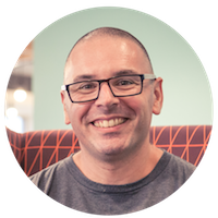 A smiling person with glasses and a shaved head, wearing a t-shirt, sitting in front of a patterned background in a circular portrait.