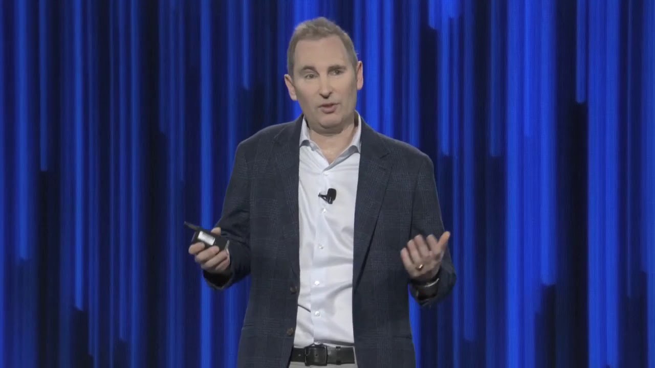 A person wearing a suit jacket and holding a clicker, standing in front of a blue stage backdrop.