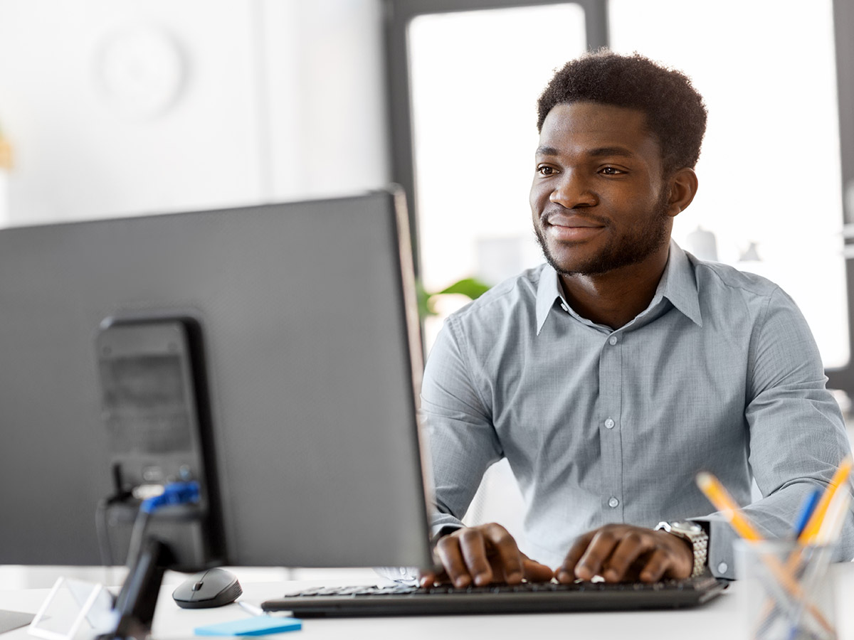 An African businessman sitting at a desk in a modern office, working on a computer and smiling.