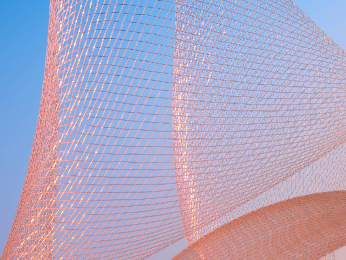 An abstract image featuring an intricate geometric wireframe or mesh structure with intersecting lines, illuminated by soft pink light, set against a clear blue sky. The visual emphasizes pattern and structure, evoking themes of analytics and data representation.