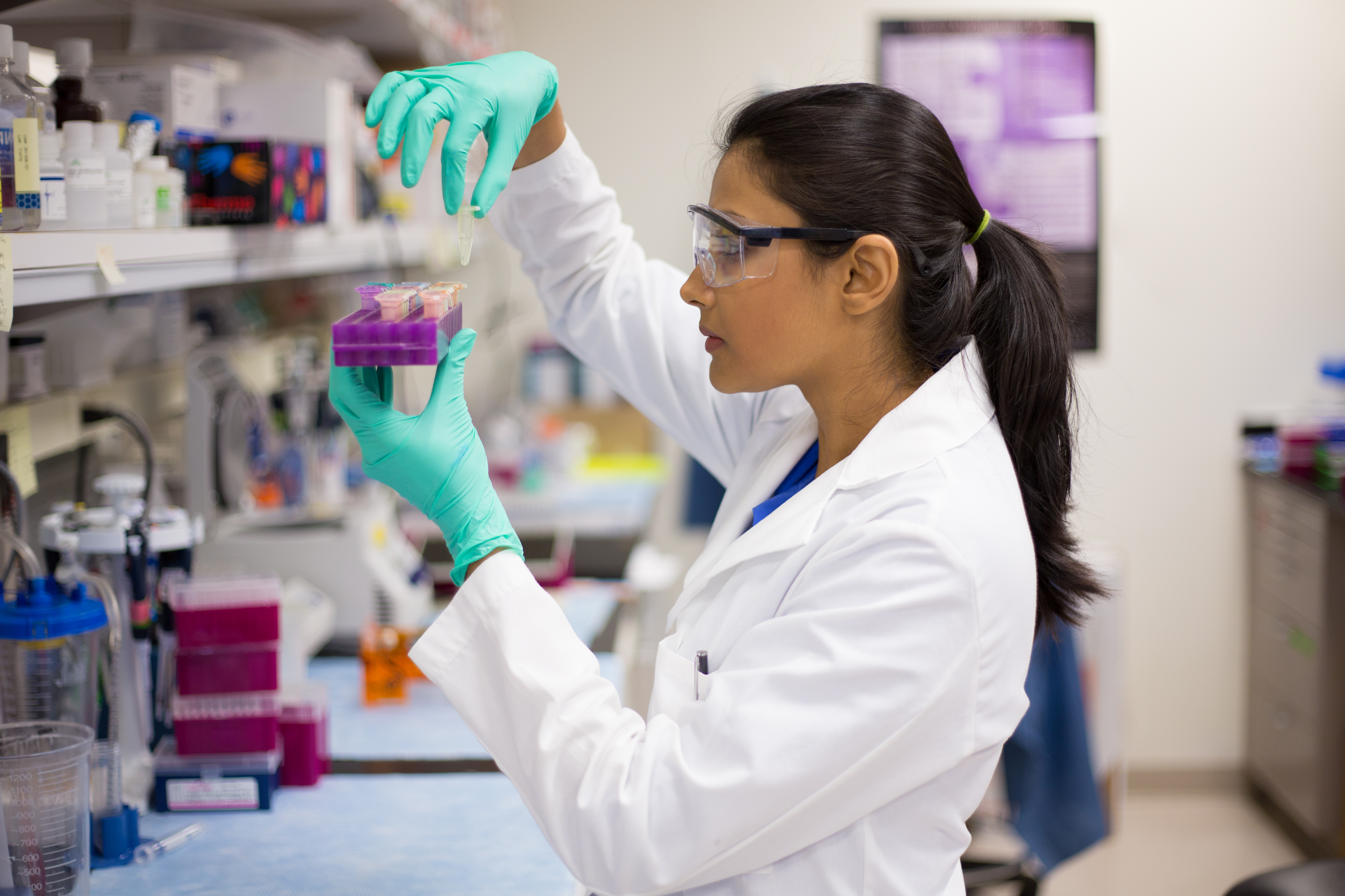 A scientist wearing safety goggles and gloves works in a laboratory, handling test tubes and colorful lab equipment for research and analysis.