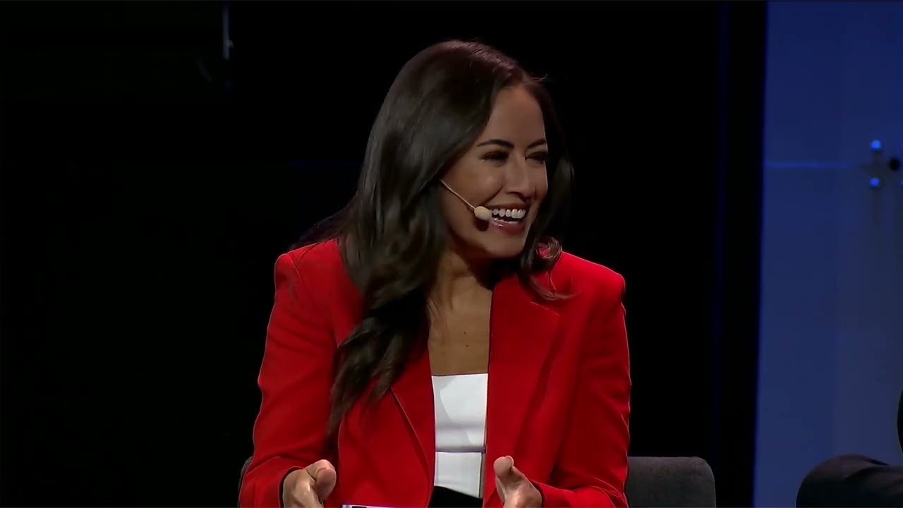 A woman wearing a red jacket and a headset microphone is speaking and smiling on stage at an event.
