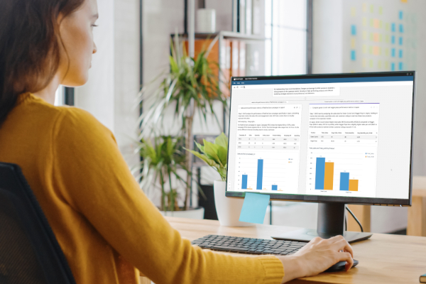 A woman sits at a desk in a modern office, analyzing bar chart data and reports displayed on a computer monitor.
